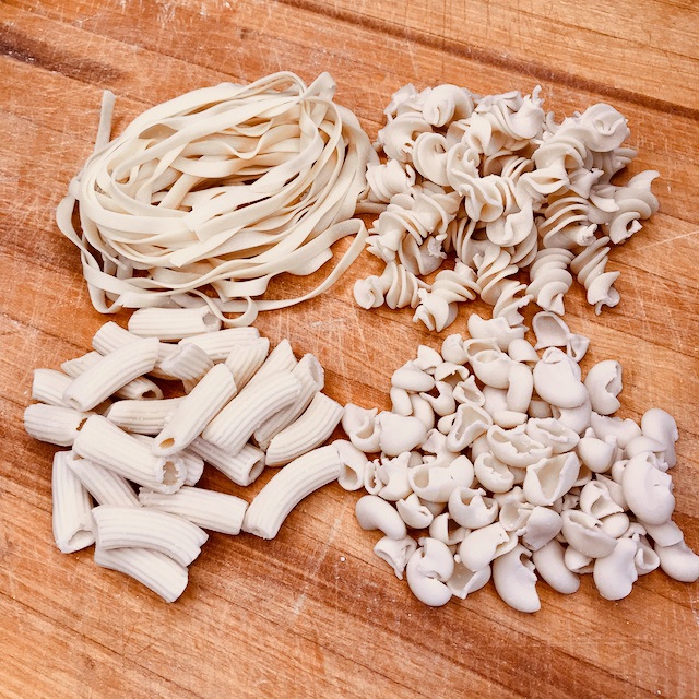 4 piles of pasta arranged in a square on a cutting board. Clockwise from top left is a small pile of tagliatelle pasta,  a small pile of fusilli pasta, a pile of shell shaped pasta and a pile of rigatoni pasta 